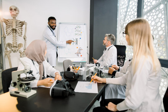 Cheerful Handsome young African doctor scientist smiling while standing near the classboard and showing new biochemical formulas for his multiracial colleagues at the meeting. Medicine, science