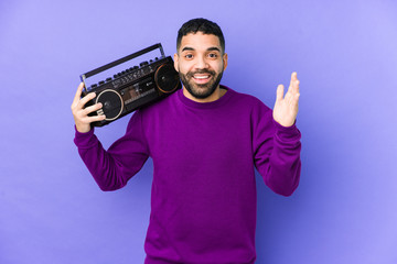 Young arabian man holding a radio cassette isolated Young arabian man listening music receiving a pleasant surprise, excited and raising hands.