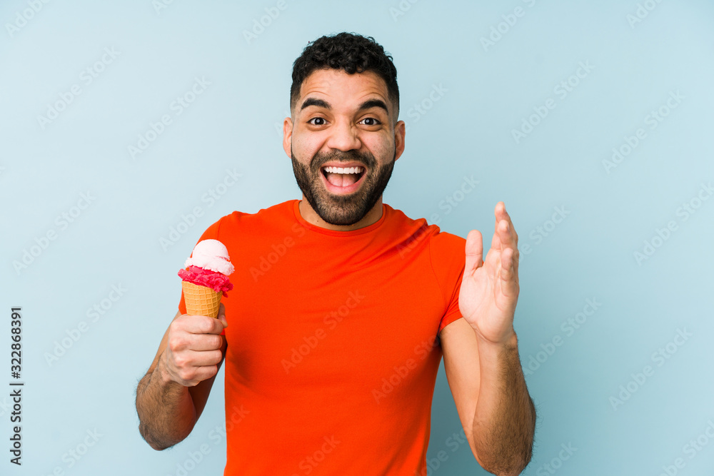 Wall mural young latin man holding an ice cream isolated receiving a pleasant surprise, excited and raising han