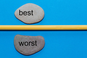 The words best and worst are written on natural smooth stones separated by a yellow pencil. The...