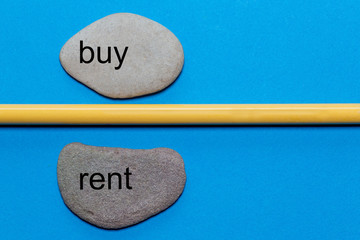 The words buy and rent are written on natural smooth stones separated by a yellow pencil. The background is isolated in blue and has a lot of space