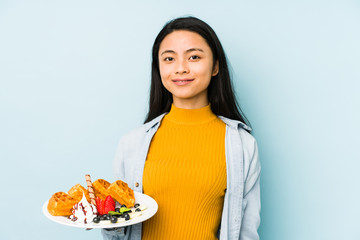 Young chinese woman holding waffle isolated on blue background  person pointing by hand to a shirt copy space, proud and confident