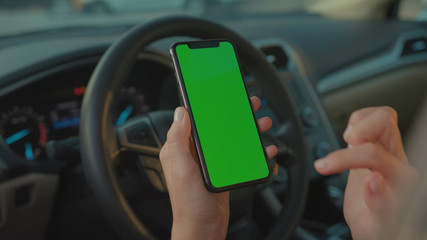 Lviv, Ukraine - May 19, 2018: Close-up of young female driver showing vertical smartphone doing tapping gestures on mock-up green screen inside modern automobile.