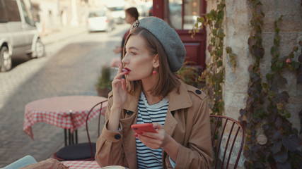 Fototapeta premium Young blonde girl woman smoking cigarette at cafe smile wearing stylish coat use phone sunny day sitting in cafe on breakfast croissant morning outdoors smile city fashion online slow motion