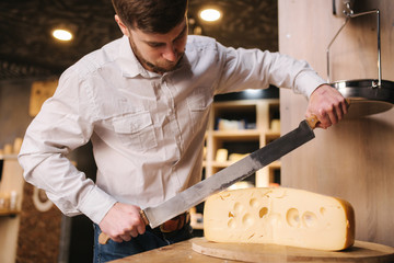 Young worker slice lactose-free cheese maasdam on wooden board in cheese shop