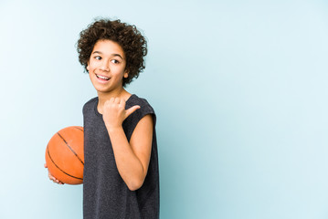 Kid boy playing basketball isolated on blue background points with thumb finger away, laughing and...