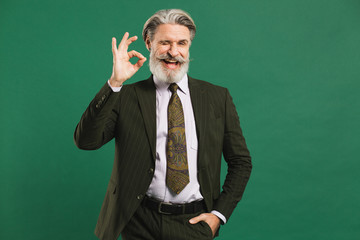 Bearded middle-aged man in khaki suit shows okay on green background with copy space.