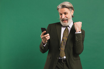 Bearded middle-aged man in khaki suit screaming loudly and looking at phone while holding fist of happiness against green background