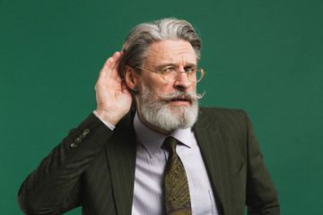 Bearded middle aged teacher in khaki suit holds ear and eavesdrop on green background
