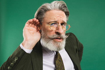 Bearded middle aged teacher in khaki suit holds ear and eavesdrop on green background