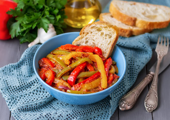 Vegetable dish Peperonata. Sauteed colorful peppers with onion, tomatoes and parsley served in blue bowl with sliced chiabatta. Wooden background, selective focus. Italian cuisine.