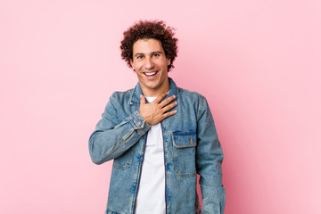 Fototapeta na wymiar Curly mature man wearing a denim jacket against pink background laughs out loudly keeping hand on chest.