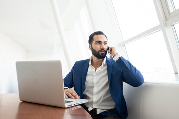 successful businessman in a suit uses a laptop and talking on the phone in a white office at the workplace