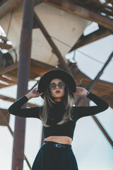 A long haired blonde woman, dressed all in black, wearing a black fedora type hat, black sun...