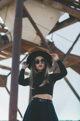 A long haired blonde woman, dressed all in black, wearing a black fedora type hat, black sun glasses, distressed industrial silo. aggregate quarry 