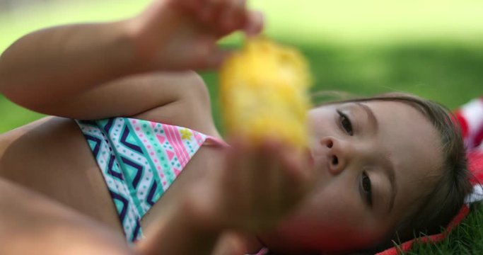 Little girl eating corn outside laid in grass. Child daydreaming while eating picnic