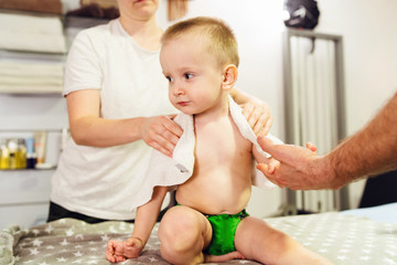 Obraz na płótnie Canvas Small caucasian child blonde son sitting on the bed while his parents mother and father are using towel to dry and clean his back on naked body after bath at home family