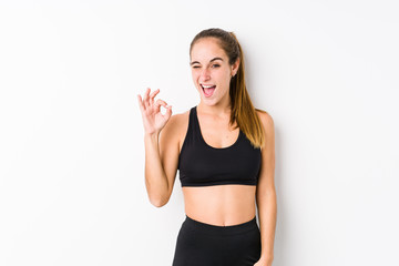 Young caucasian fitness woman posing in a white background winks an eye and holds an okay gesture with hand.
