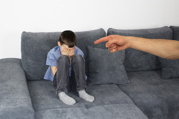 sad child, a boy sitting on a gray sofa in the room and covered his face with his hands, the hand...