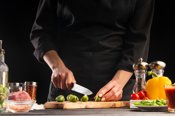 Chef slices Brussels sprouts on the background of vegetable ingredients, on a black background. Cooking salad. Healthy and wholesome food, cuisine and cooking, recipe book.