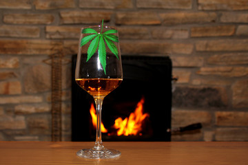 one glass of wine and a leaf of marijuana stands on a table in front of a fireplace in which firewood burns with a bright flame