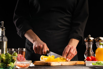 Chef slices sweet orange bell pepper on the background of vegetable ingredients, on a black background. Cooking salad. Healthy and wholesome food, cuisine and cooking, recipe book.