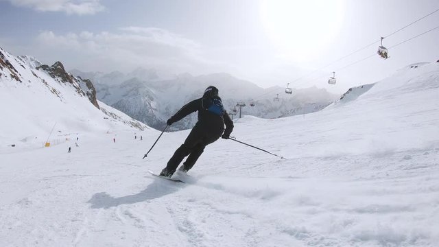 Slow motion shot of man skiing next to the ski lift In Ponte di Legno. He is wearing a bag. There are beautiful mountains.