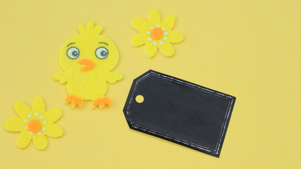 yellow felt crafted chick and flowers for easter next to a        black blank tag with writing space