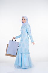 A beautiful Muslim female model in a Asian traditional dress modern kurung carrying shopping bags isolated on white background. Eidul fitri festive preparation shopping concept. Full length portrait.