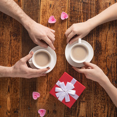 Valentine's Day celebration concept. A nice gift for your loved one. Hands of man and woman with coffee mugs on a wooden table background. Copy space. Flat lay. Close-up.