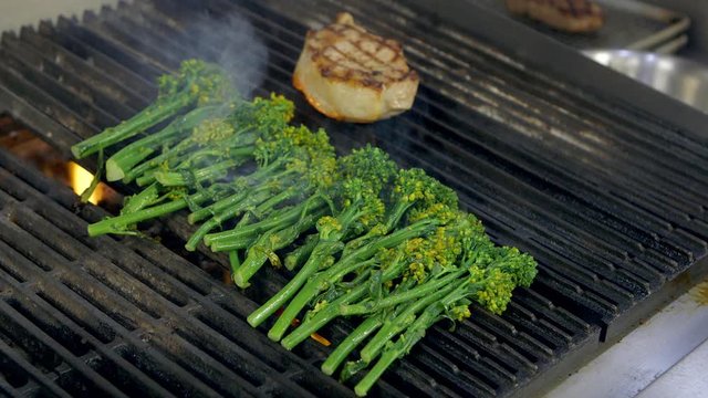 Fresh broccolini and a pork chop cook on a grill in a restaurant kitchen.