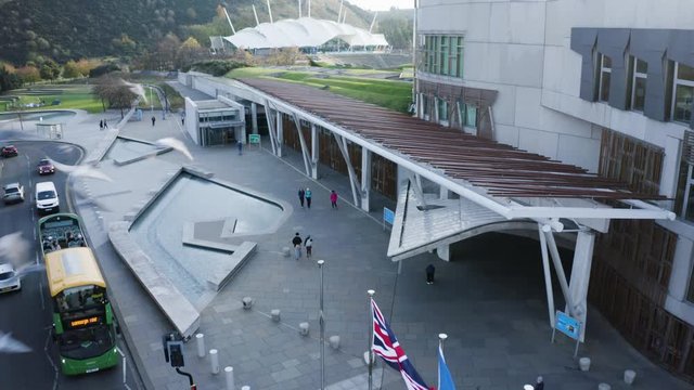 A low shot above the road in front of the Holyrood Scottish Parliament, with a tourist bus, traffic and tourists. Union jack, flag, Scottish flag, EU flag flying | Edinburgh, Scotland | 4K at 30fps