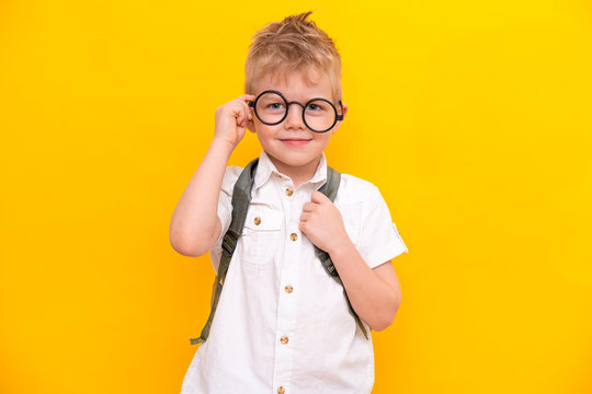 Back to school. Portrait of funny clever blonde school boy in round glasses with bag in white shirt. Yellow studio background. Education. Looking and smiling at camera