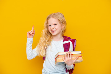 Back to school. Portrait of blonde school girl with bag and books. Pointing finger up. Yellow studio background. Education. smiling at camera - obrazy, fototapety, plakaty
