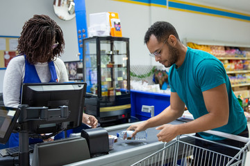 African American cashier scanning goods at counter. Tranquil bearded man putting products on...
