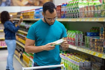 Focused African American man reading information on packaging. Concentrated bearded guy buying food at supermarket. Shopping concept