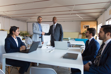 Two businessmen shaking hands in office. Smiling colleagues standing and looking at camera. Business meeting concept
