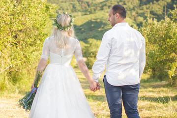 Fototapeta na wymiar Back view on bride and groom holding hands in sunny summer day. Outdoor wedding and relationsheep romantic concept