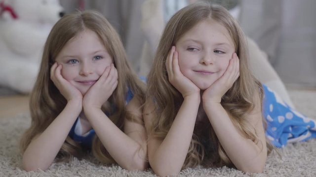 Close-up of happy Caucasian twin sisters looking at each other and turning to camera as lying and holding head with hands. Brunette girls with long wavy hair resting at home. Lifestyle, family.