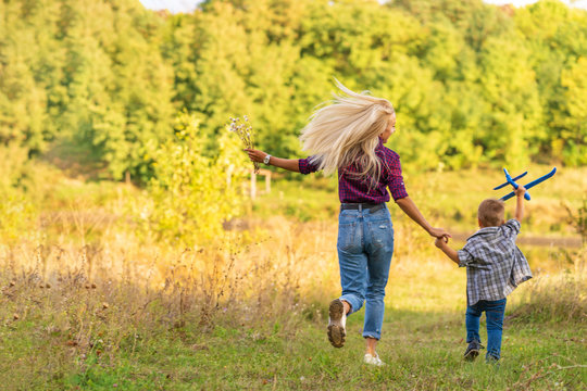 Little Boy Playing With Toy Airplane With His Young Mom Outdoors At Sunset. Happy Kid Is Playing In Park Outdoors.