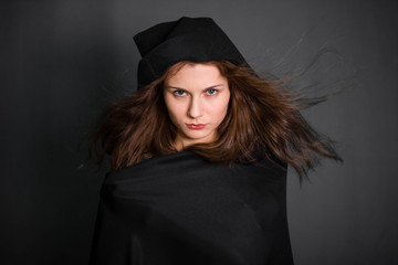 Young, beautiful girl in a black cloak with a hood in the studio on a gray background. Long hair fluttering on the wind. A model with clean skin. Suit of a fortuneteller, witches for Halloween.