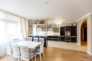 Large Kitchen-living room in the cottage with kitchen and dining area. Luxury style. The kitchen is black and white, and the dining table and chairs are white. 