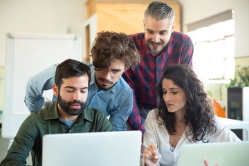 Fototapeta premium Confident young coworkers looking at laptop. Thoughtful male and female coworkers standing and sitting at table with laptops in open space office and communicating. Startup, teamwork concept