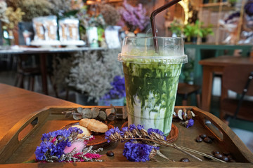 Close up plastic take away cup of iced Matcha green milk tea served with butter cookies on wooden tray table decorated with dry flower and coffee beans