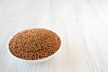 Uncooked Roasted Buckwheat in a white bowl on a white wooden surface, low angle view. Copy space.