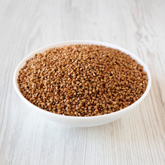 Uncooked Roasted Buckwheat in a white bowl on a white wooden table, side view.