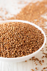 Uncooked Roasted Buckwheat in a white bowl on a white wooden surface, side view. Close-up.