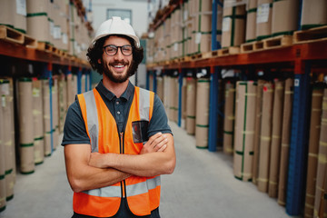 Cheerful male worker entrepreneur in uniform with white hardhat and arms crossed standing in from of warehouse rack arrangement wearing spectacles