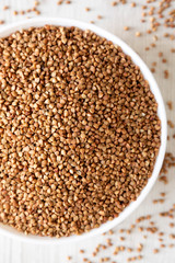 Uncooked Roasted Buckwheat in a white bowl on a white wooden table, top view. Flat lay, overhead, from above. Close-up.