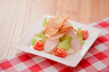 Plate of traditional Caesar Salad with chicken and salad on light wooden table with red plaid tablecloth.